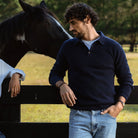 A man wearing a navy blue knit polo sweater and light blue jeans stands outdoors next to a black fence, with a horse visible behind him. The product being shown is the navy sweater.