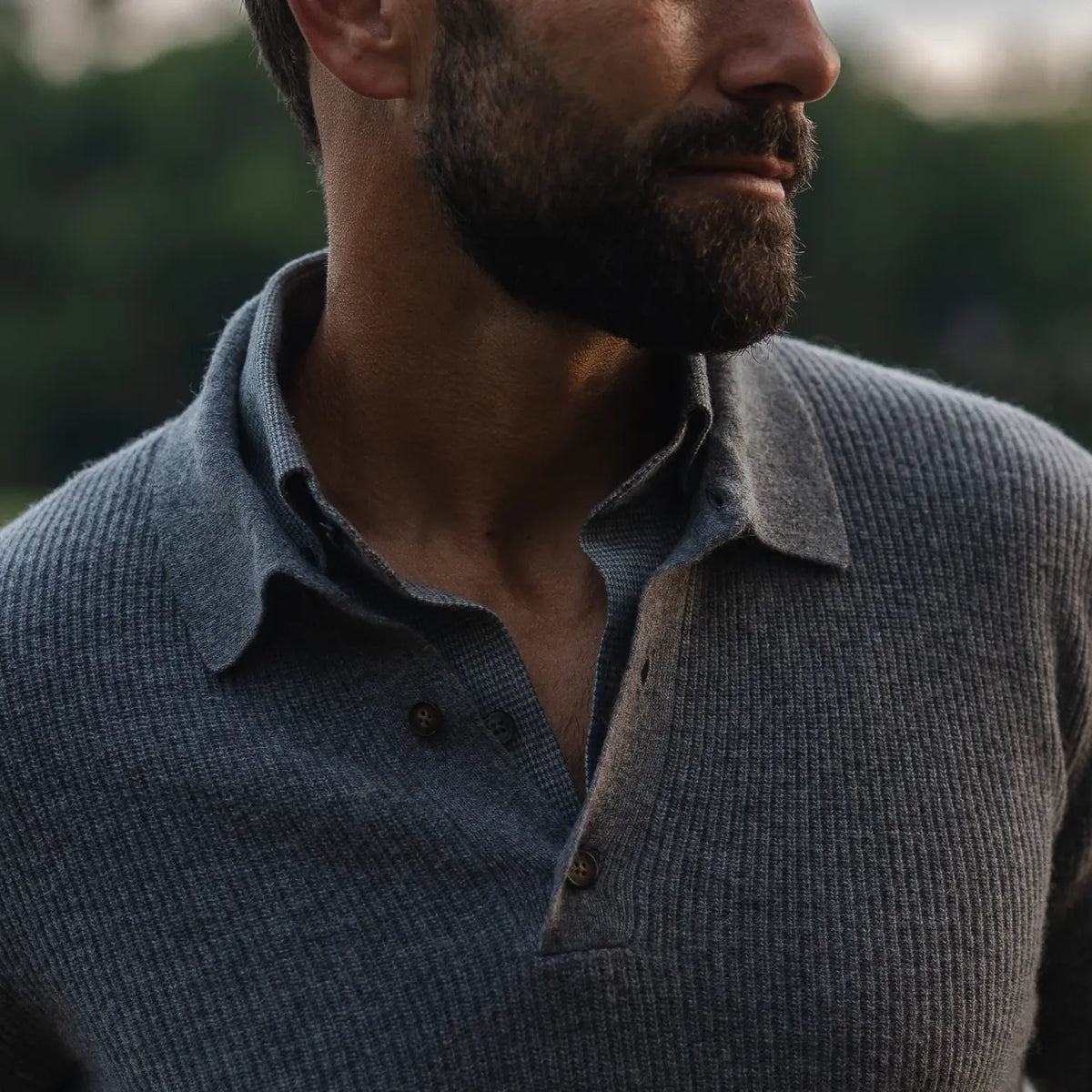 Close-up of a man wearing a textured gray knit polo neck sweater outdoors, with a blurred natural background.