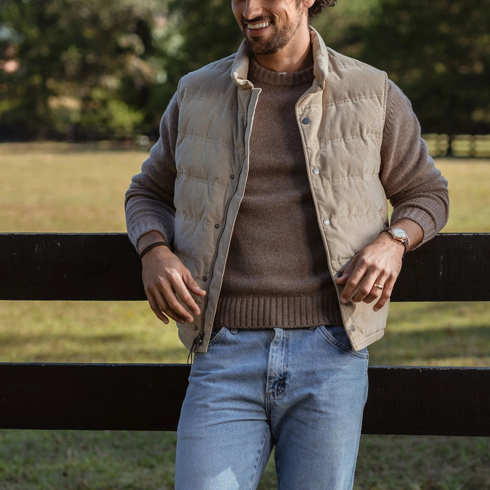 A man wearing a beige quilted puffer vest over a brown knit sweater and light blue jeans, standing outdoors in front of a dark wooden fence with a blurred green background.