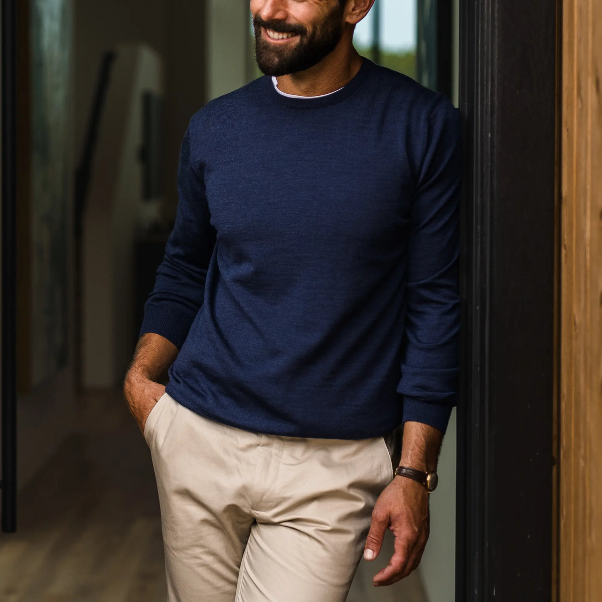 A man wearing a navy blue long-sleeve crewneck sweater and beige trousers, leaning against a dark doorway in an indoor setting.