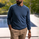A man wearing a heathered blue long-sleeve crewneck sweater and khaki pants, standing outdoors near a pool with a blurred green background.