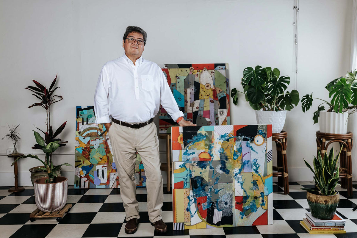 Man standing in front of colorful abstract paintings in a room with plants and checkered floor.