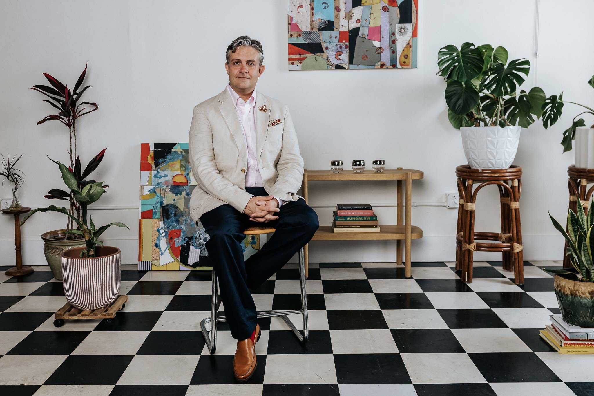 Man sitting on a chair in a room with checkered floor, plants, and colorful artwork.