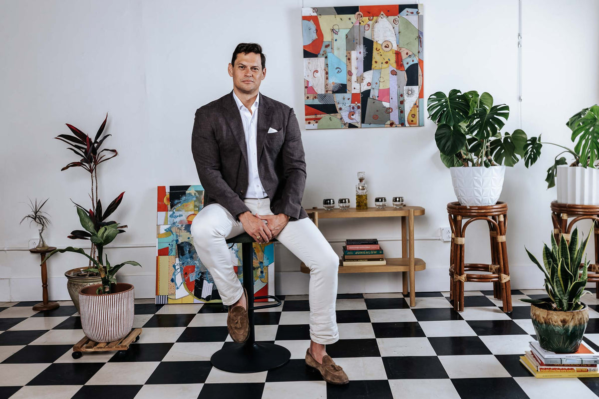 Man sitting on a stool in a room with checkered floor, plants, and colorful artwork.
