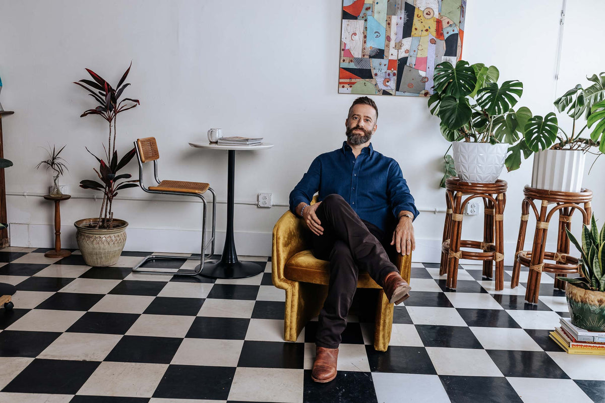 Man sitting in a chair in a room with checkered floor, plants, and a colorful wall art piece.