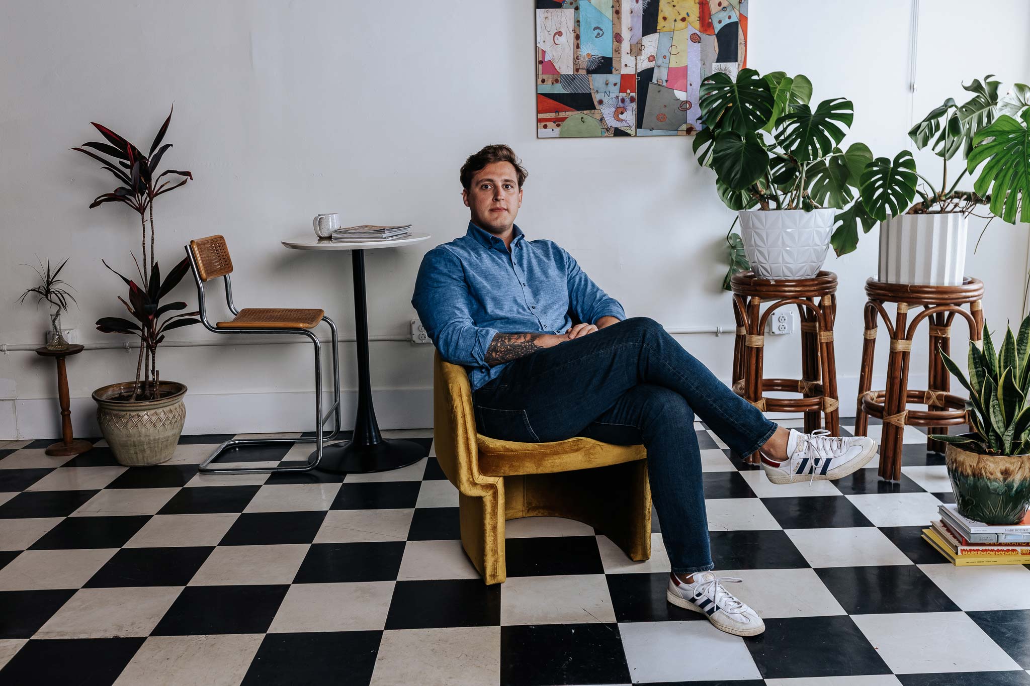 Man sitting on a yellow chair in a room with a checkered floor, plants, and a colorful abstract painting.