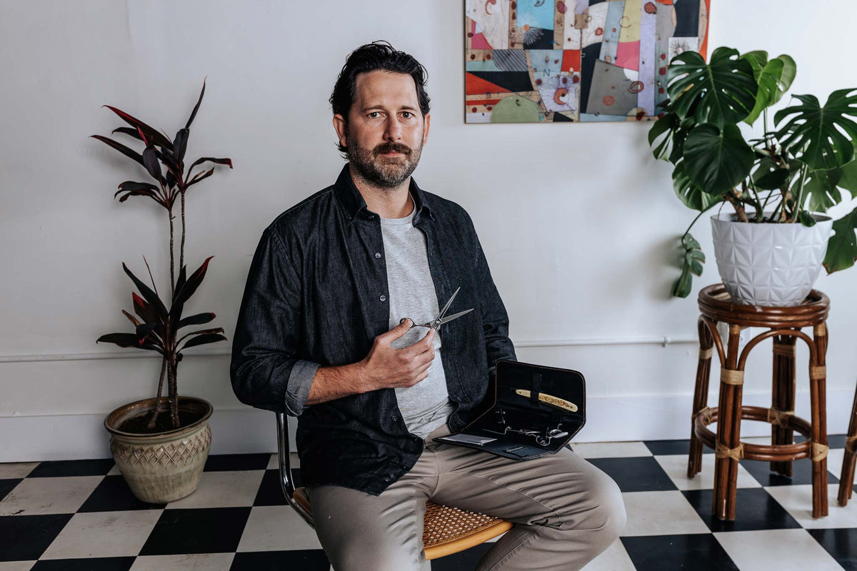 Man sitting in a chair holding scissors in a room with plants and checkered floor.