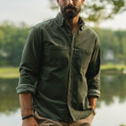 A man wearing an olive green long-sleeve button-up shirt with two chest pockets, standing outdoors near a body of water with trees in the background.