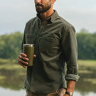 A man outdoors by a lake wearing an olive green long-sleeve button-up shirt and holding an olive green insulated tumbler.