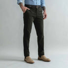 A man wearing slim-fit dark olive green corduroy trousers, styled with a black belt, a light blue shirt, and tan suede boots, standing against a light gray studio background.