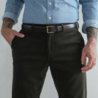 A close-up of a man wearing dark olive green corduroy trousers, a dark brown leather belt with a gold buckle, and a light blue button-down shirt. His hands are in his pockets, and he has visible tattoos on his arms.