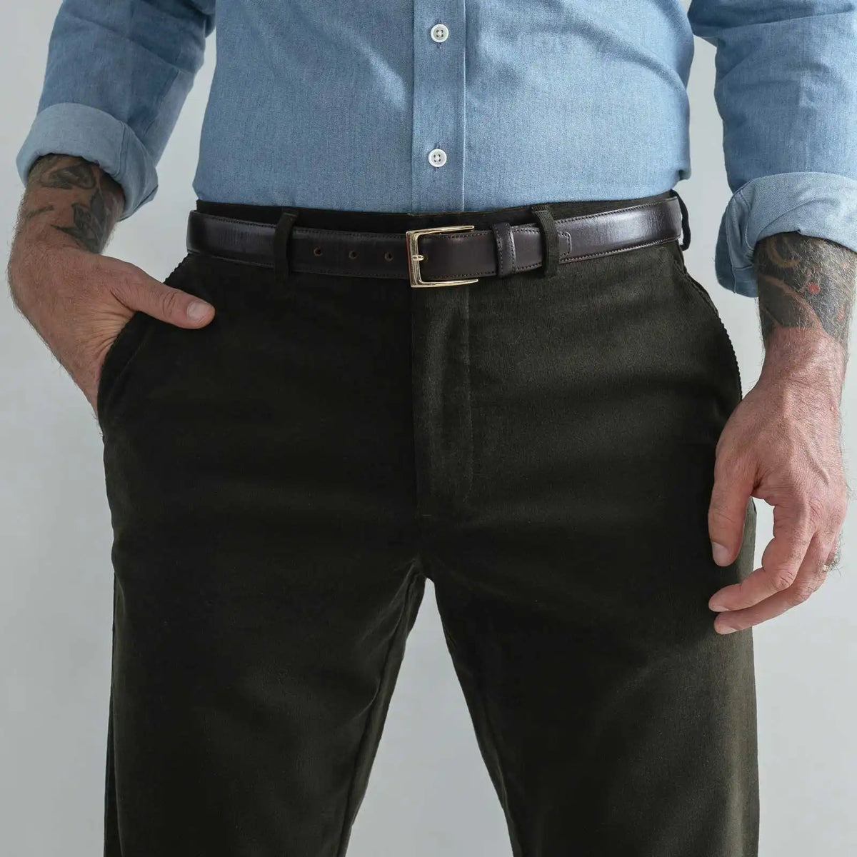 A close-up of a man wearing dark olive green corduroy trousers, a dark brown leather belt with a gold buckle, and a light blue button-down shirt. His hands are in his pockets, and he has visible tattoos on his arms.