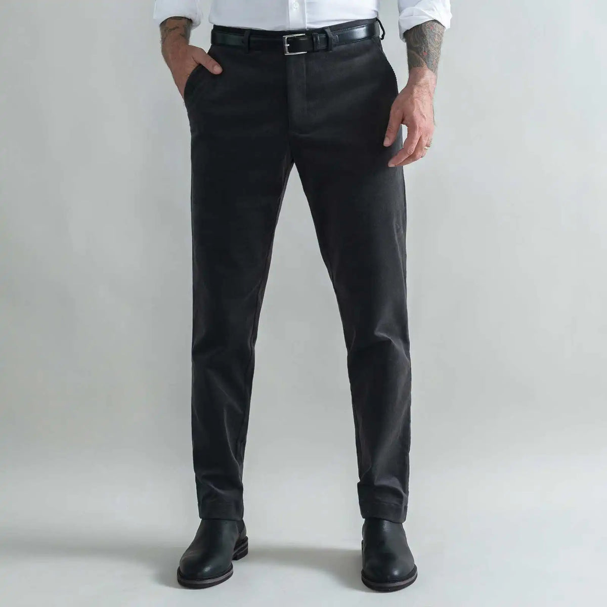 A man wearing slim-fit dark grey corduroy trousers, a black belt, a white shirt, and black leather boots, standing against a light gray studio background.