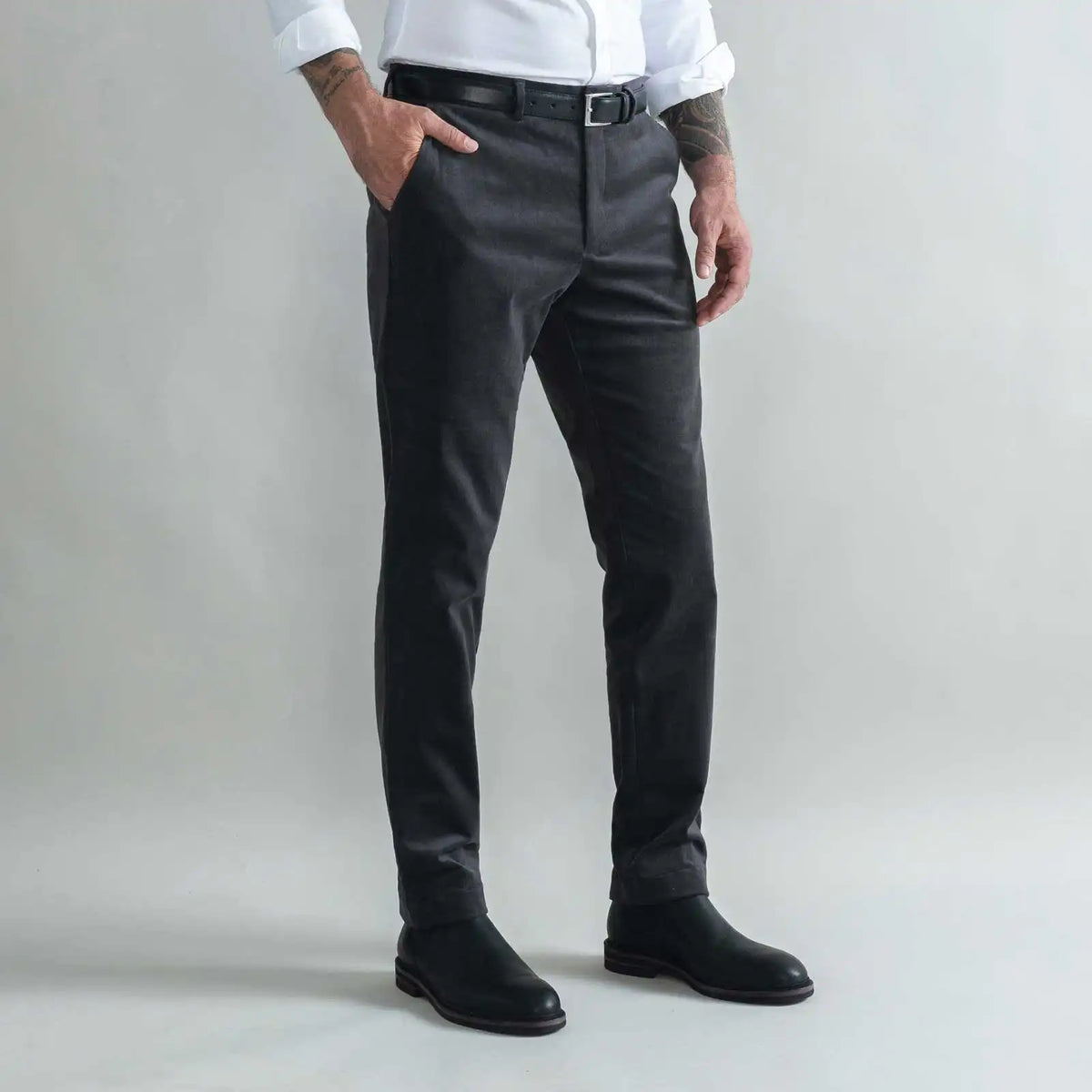 A man wearing slim-fit charcoal gray corduroy chinos, styled with a white shirt, black belt, and black boots, standing against a light gray studio background.