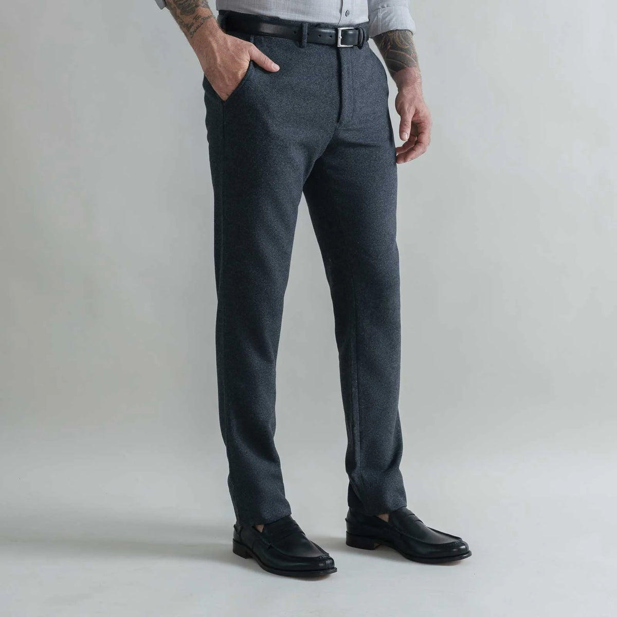 A man wearing dark gray heathered wool trousers, a black belt, a light gray shirt, and black leather loafers, standing against a light gray studio background.