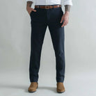 A man wearing dark navy blue slim-fit trousers, styled with a brown leather belt, a white dress shirt, and brown suede boots, standing against a light gray studio background.