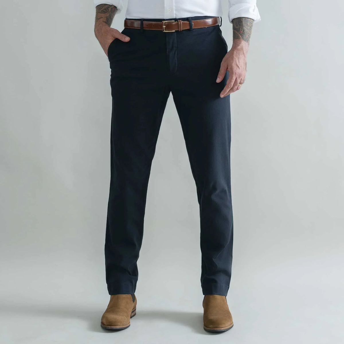 A man wearing dark navy blue slim-fit trousers, styled with a brown leather belt, a white dress shirt, and brown suede boots, standing against a light gray studio background.