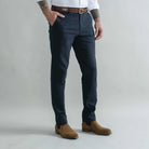 A man wearing navy blue slim-fit trousers, styled with a brown leather belt, a white shirt, and brown suede Chelsea boots, standing against a light gray studio background.