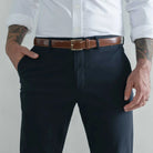 A man wearing dark navy trousers, a brown leather belt with a gold buckle, a white button-down shirt, and tattoos on both arms, standing against a light gray studio background.