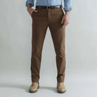 A person wearing slim-fit brown trousers, a black belt, a light blue button-up shirt, and tan boots, standing against a plain light gray studio background.