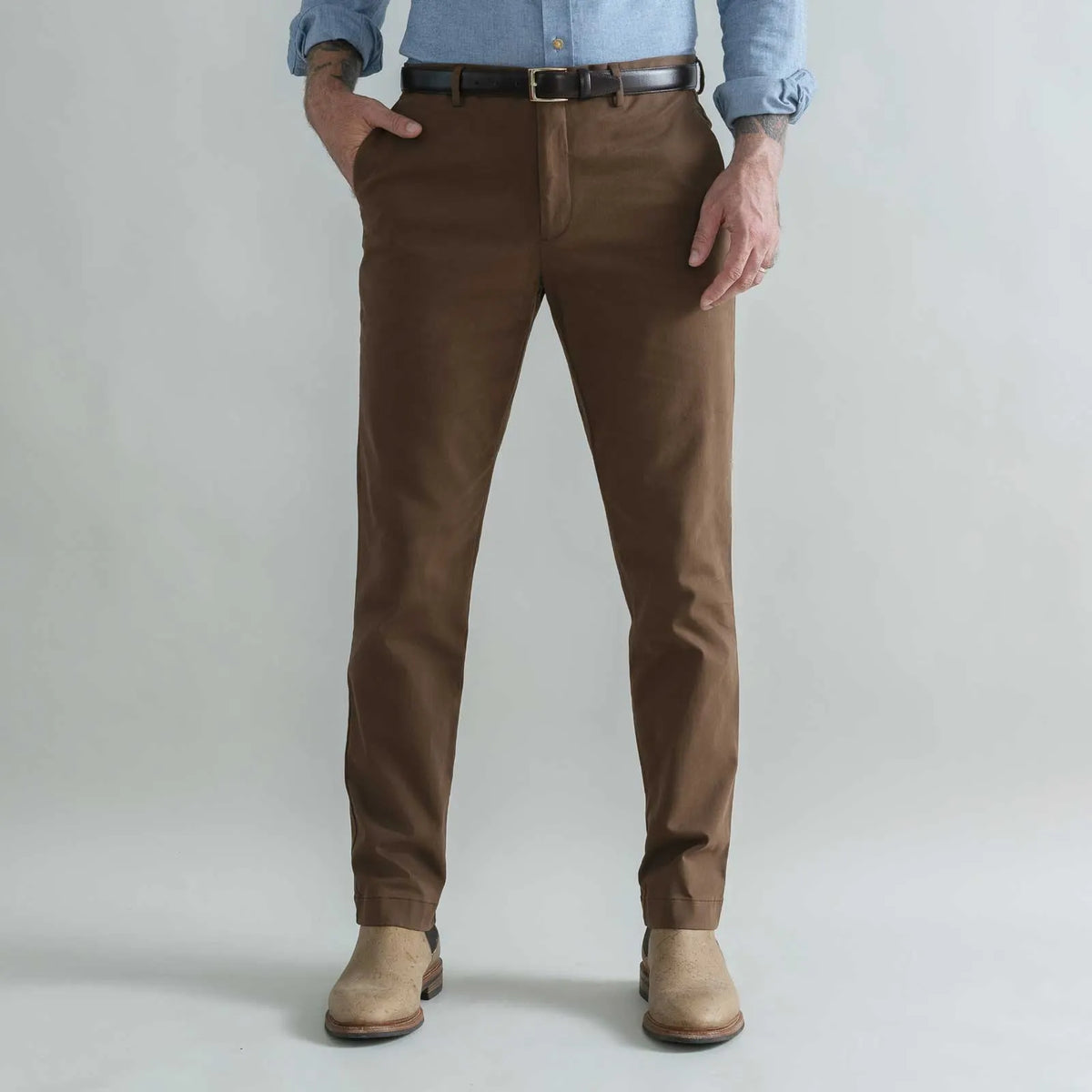 A person wearing slim-fit brown trousers, a black belt, a light blue button-up shirt, and tan boots, standing against a plain light gray studio background.