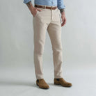 A man wearing tan moleskin chinos with a brown belt, a light blue shirt, and brown suede boots, standing against a light gray studio background.