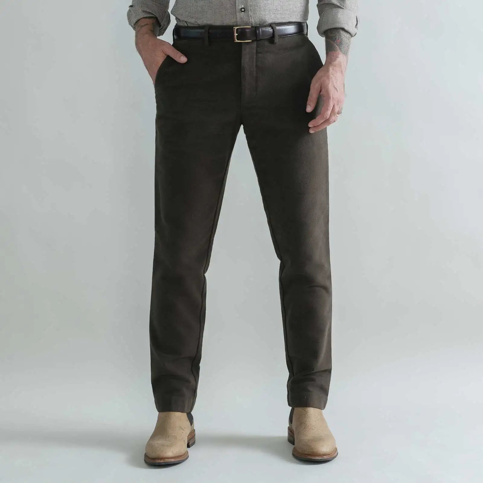 A person wearing slim-fit dark brown moleskin trousers, styled with a black belt, a gray shirt, and beige boots, standing against a light gray studio background
