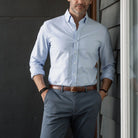 Man wearing a light blue Oxford dress shirt and gray pants against a dark wall.