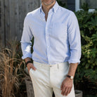 Man wearing a light blue striped Mayfield Oxford shirt and white pants standing outdoors.
