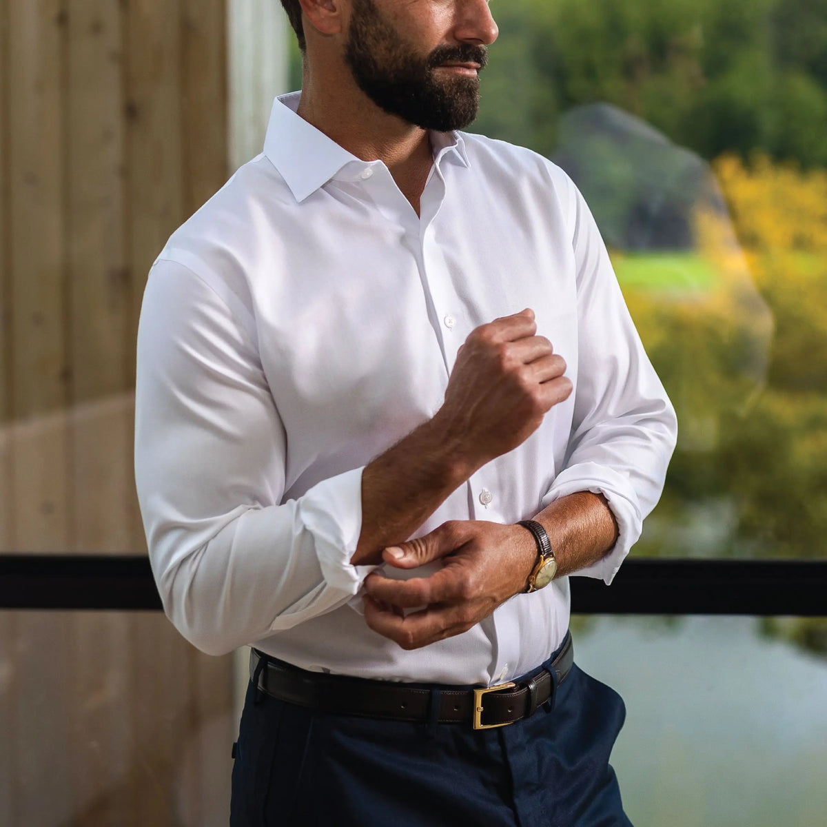 A man wearing a crisp white long-sleeve button-down dress shirt, dark navy trousers, and a black belt, standing by a window with a blurred outdoor background.