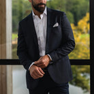 A man wearing a dark navy blue suit jacket over a white dress shirt, standing in front of a window with a blurred outdoor background.
