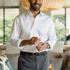 A man wearing a crisp white long-sleeve button-down dress shirt, dark gray trousers, and a black belt with a gold buckle, standing in a modern living room setting.