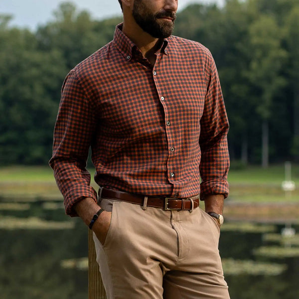A man wearing a rust-orange and brown checkered long-sleeve button-down shirt, khaki pants, and a brown belt, standing outdoors near a lake with trees in the background.