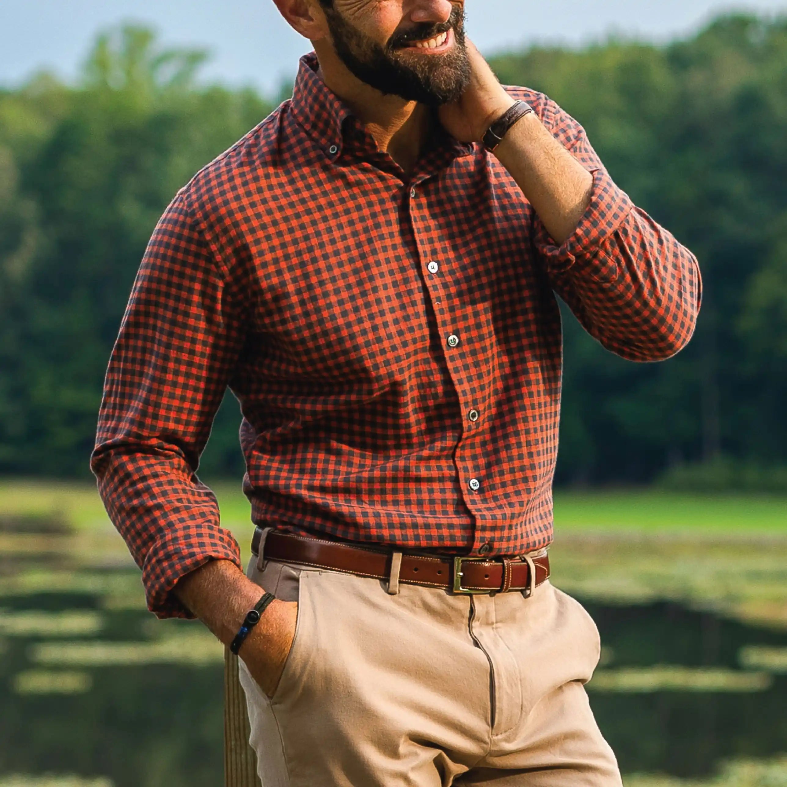 A man wearing an orange and black checkered long-sleeve button-down shirt, khaki pants, and a brown belt, standing outdoors in front of a blurred green background.