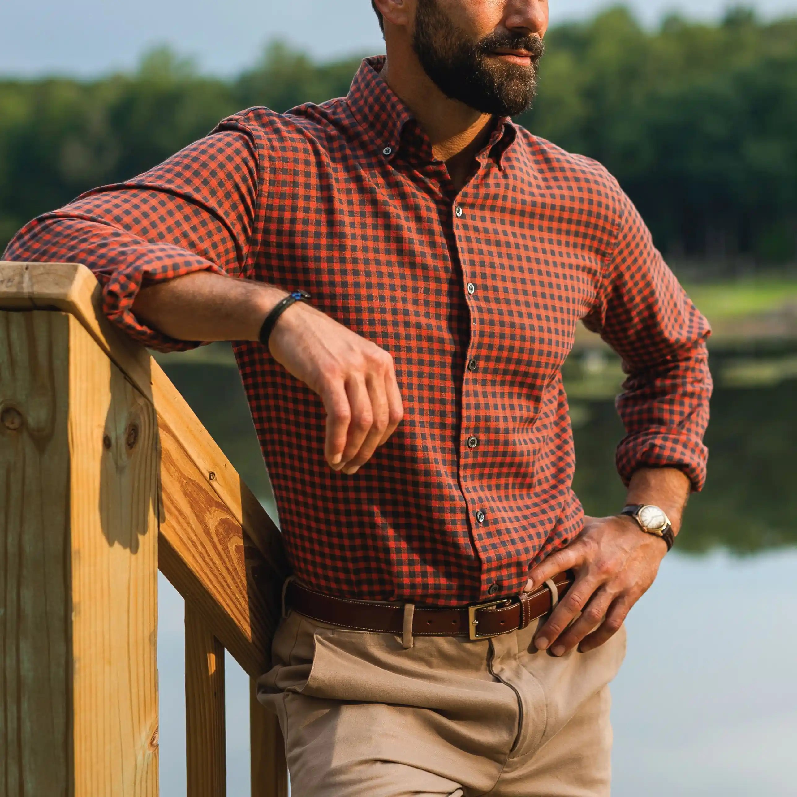 A man wearing an orange and black checkered long-sleeve button-down shirt, khaki pants, and a brown leather belt, leaning on a wooden railing outdoors near a lake.