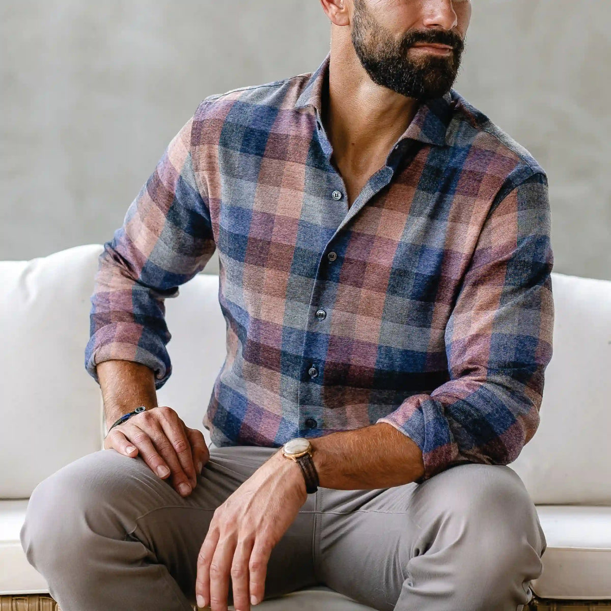 A man wearing a long-sleeve button-down plaid shirt in muted blue, pink, and beige tones, paired with light gray trousers, sitting on a white couch.