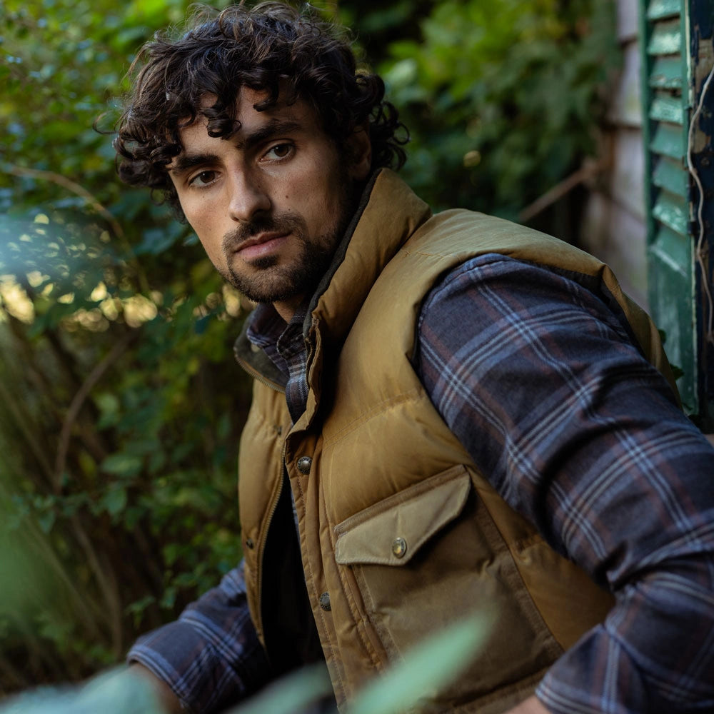 A man wearing a tan quilted puffer vest over a dark blue and gray plaid flannel shirt, photographed outdoors against a blurred green garden background.