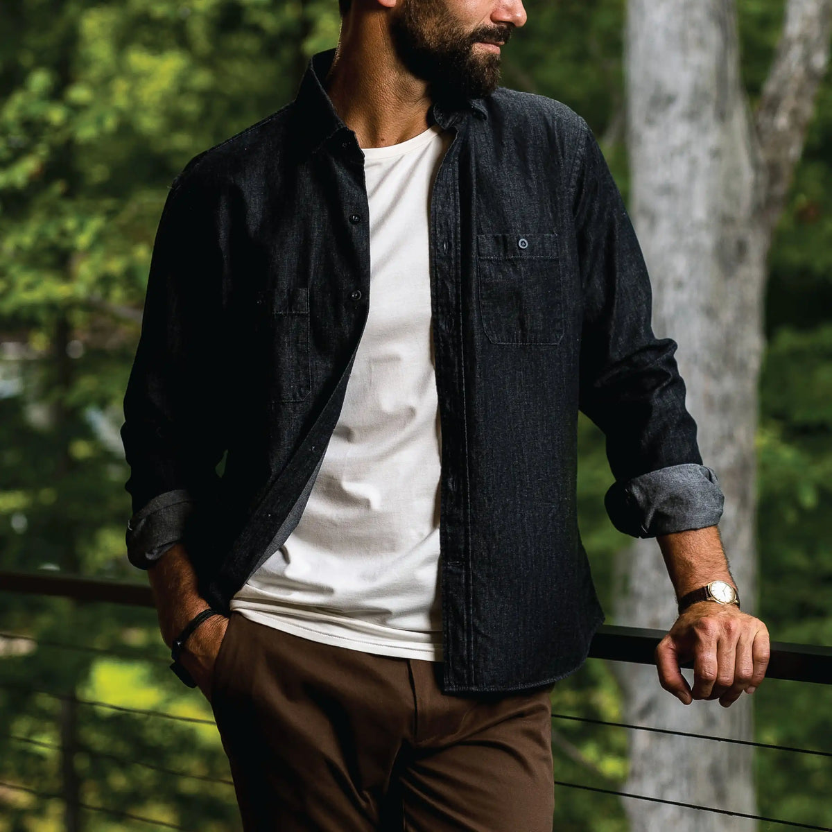 A man wearing a dark charcoal denim button-up shirt over a white t-shirt and brown pants, leaning on a railing in an outdoor forest setting.