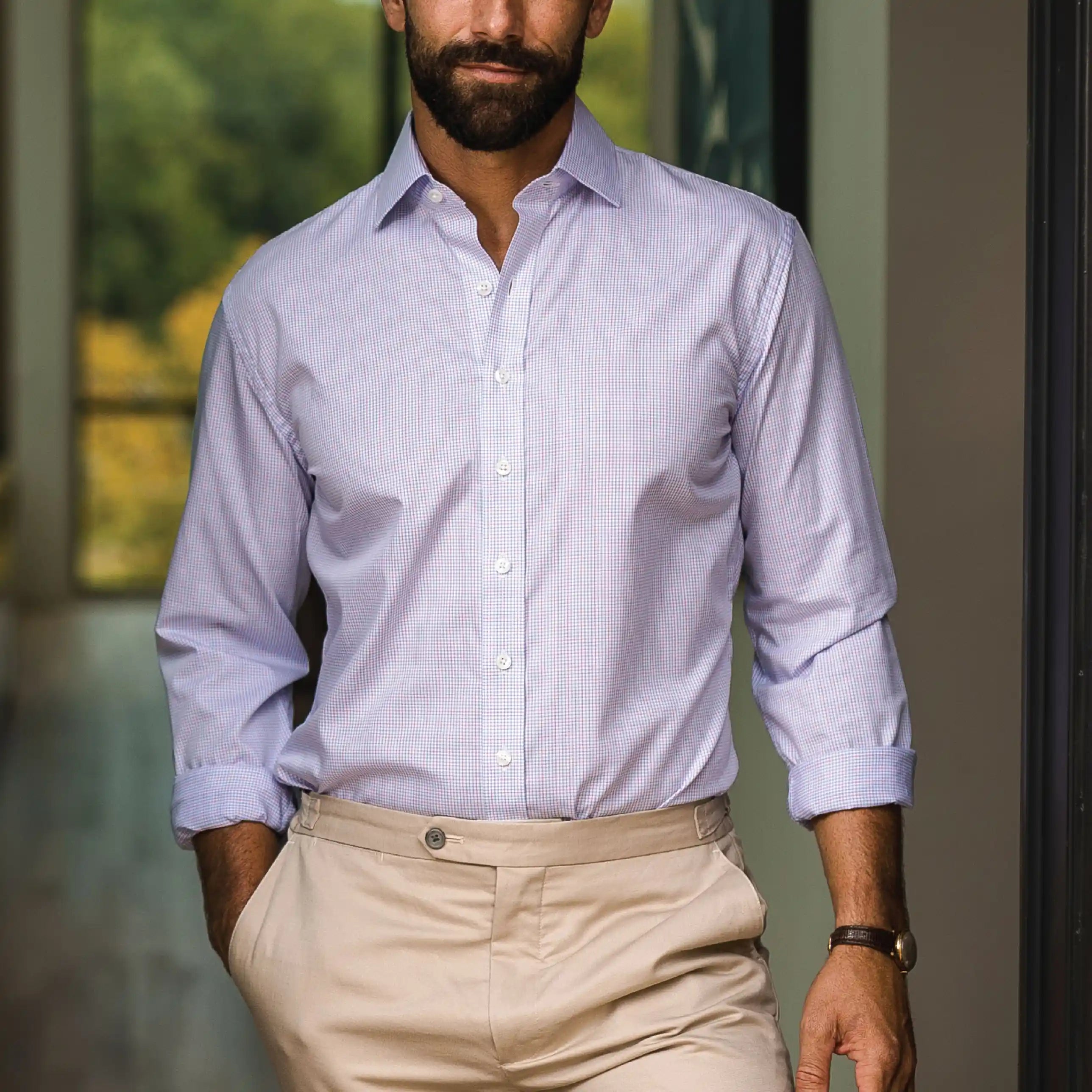 A man wearing a light pink long-sleeve button-down dress shirt with beige trousers, standing in front of a softly blurred modern interior background.