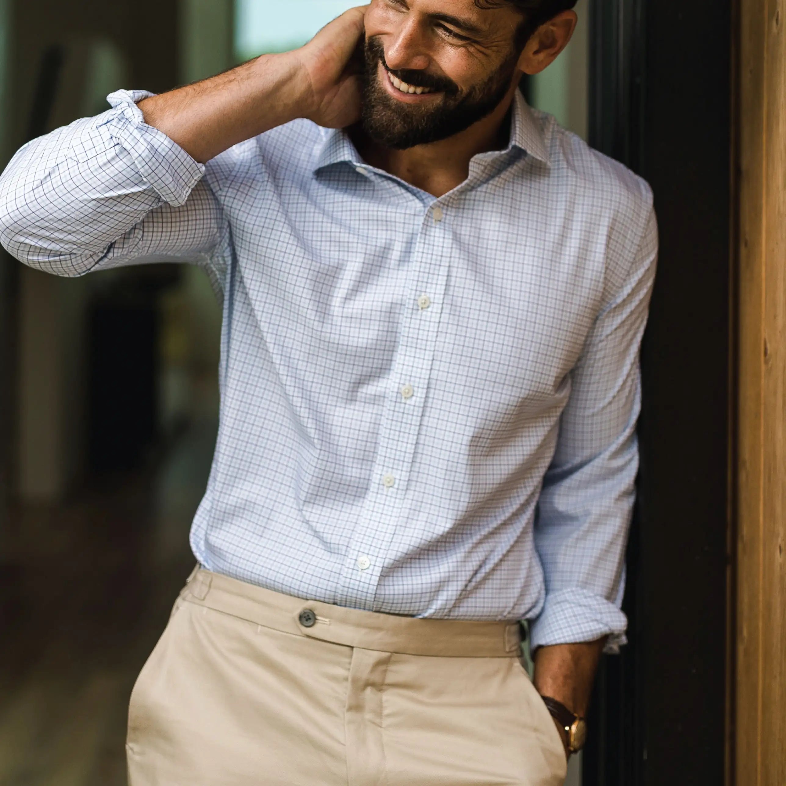 A man wearing a light blue and white checkered long-sleeve button-down shirt tucked into beige trousers, smiling and resting his head on his hand.