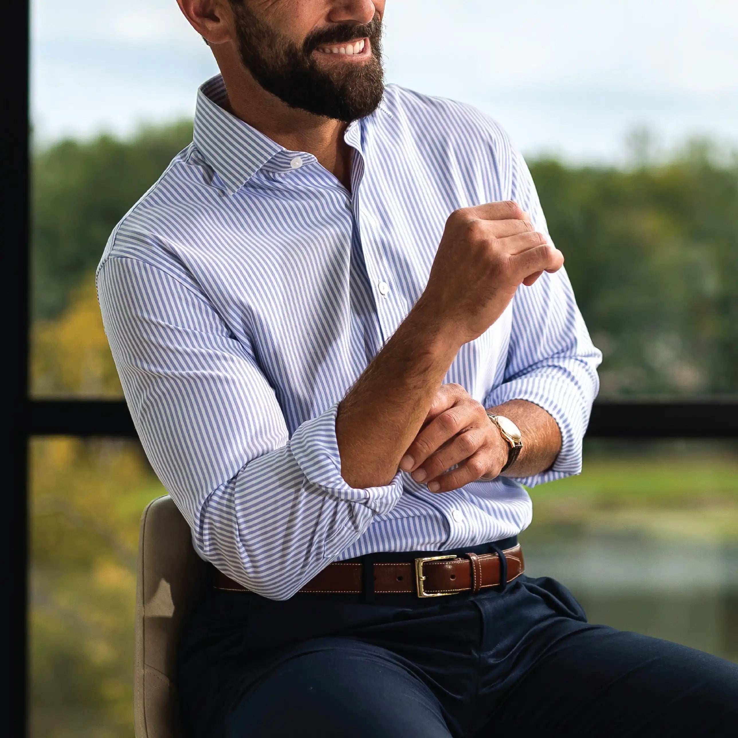 A man wearing a light blue and white striped button-down shirt, dark navy trousers, and a brown leather belt, sitting in a chair with a blurred outdoor background.