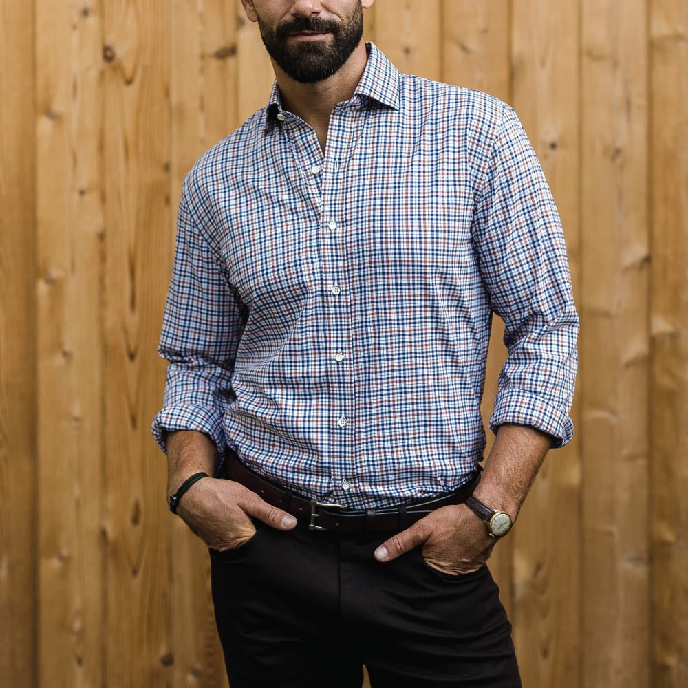 A man wearing a blue, white, and dark brown plaid button-down shirt with black trousers, standing in front of a wooden fence.