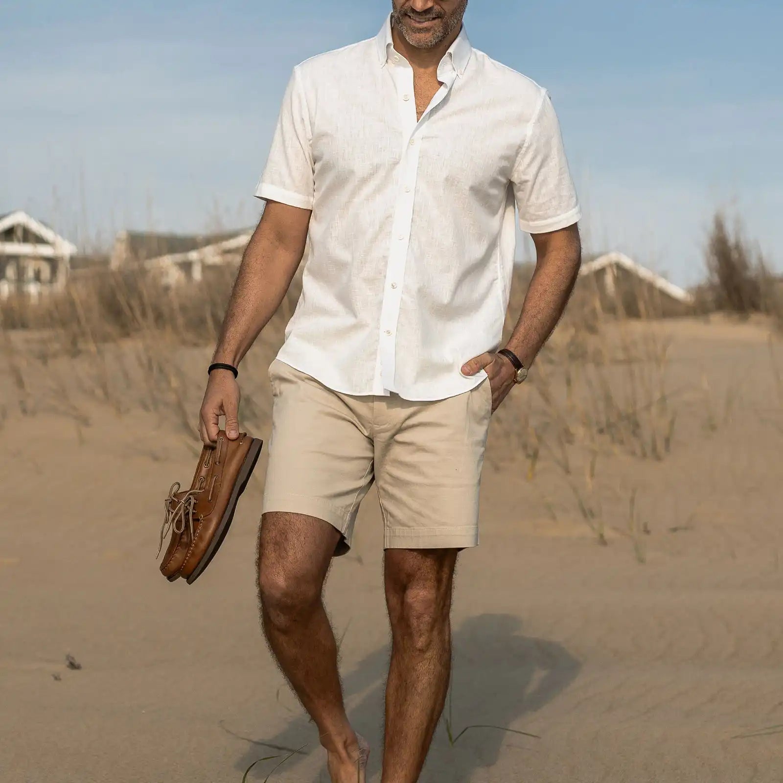 Man walking on a sandy beach wearing a white shirt and beige shorts, holding brown shoes.
