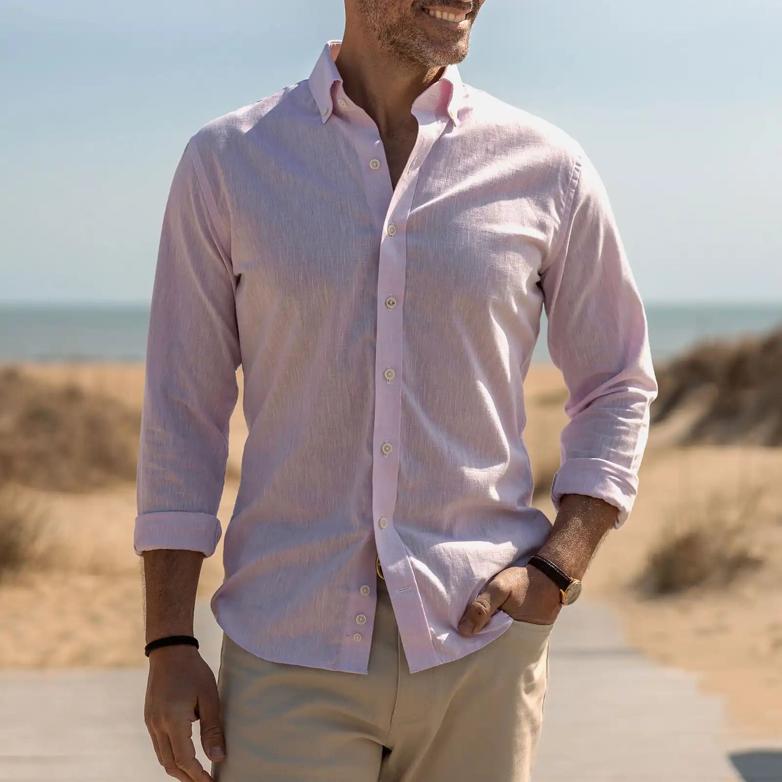 Man wearing a light pink button-up shirt and beige pants standing on a beach.