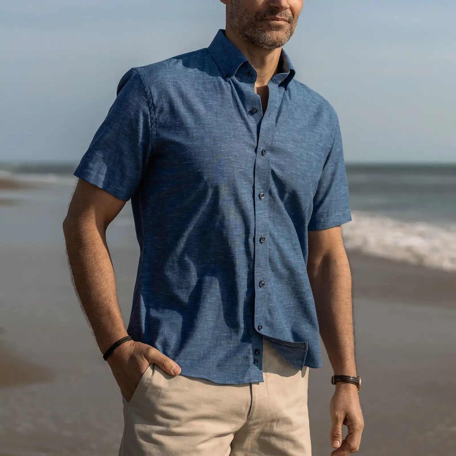 Man wearing a short sleeve blue cotton button down shirt and beige pants standing on a beach.