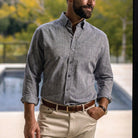 A man wearing a gray long-sleeve linen button-down shirt, paired with beige trousers and a brown belt, standing in front of a blurred outdoor background.
