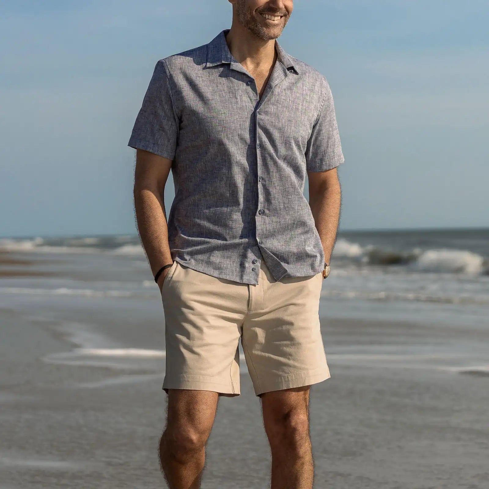 Man standing on a beach wearing a blue camp collar cotton shirt and beige shorts.