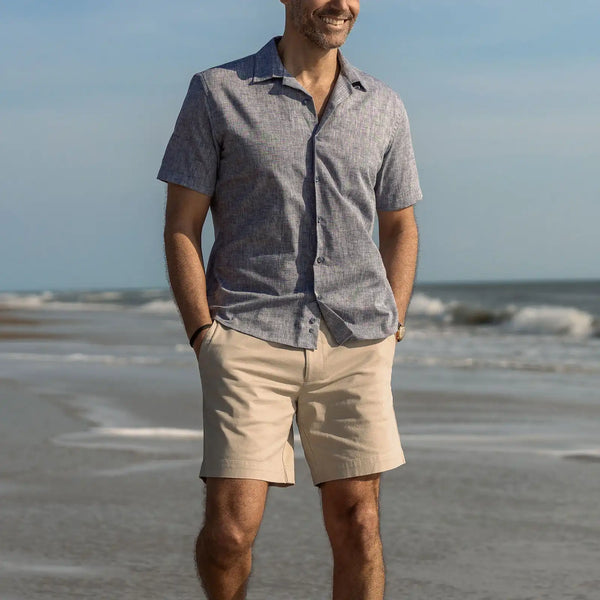 Man standing on a beach wearing a blue camp collar cotton shirt and beige shorts.