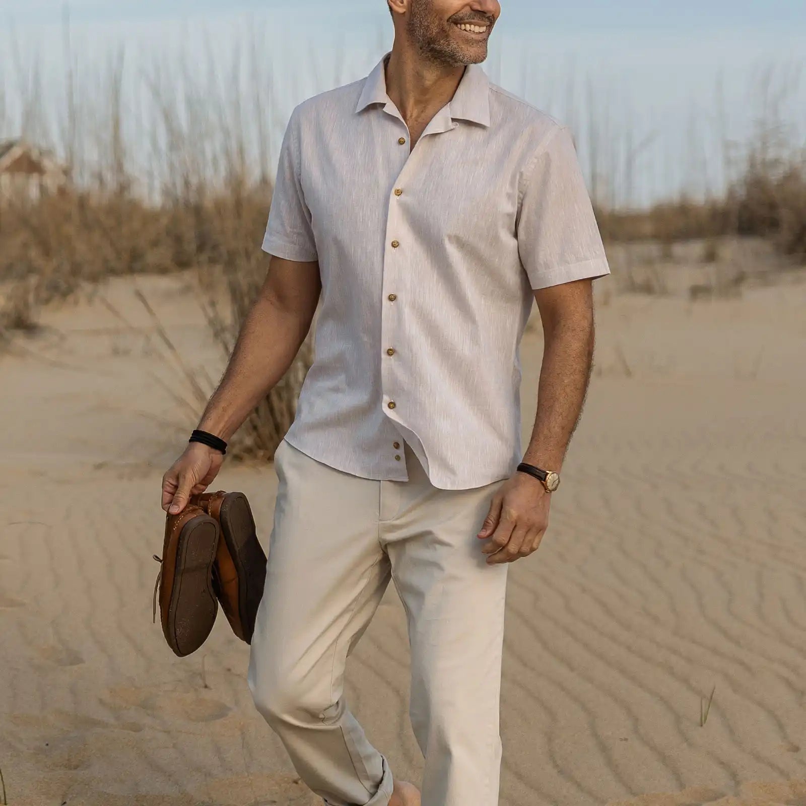 Man in a cream colored camp collar button down shirt and khakis, holding shoes, in a beach setting