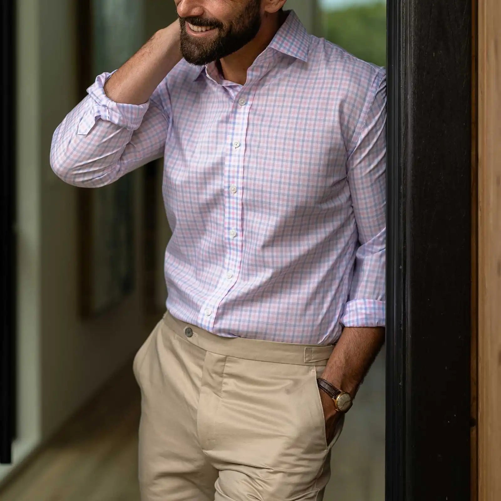 A man wearing a light pink, light blue, and white checkered button-down shirt with beige trousers, standing in a doorway.